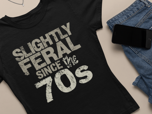 Black t-shirt with 'Slightly Feral Since the 70s' text on a beige surface with jeans and phone.
