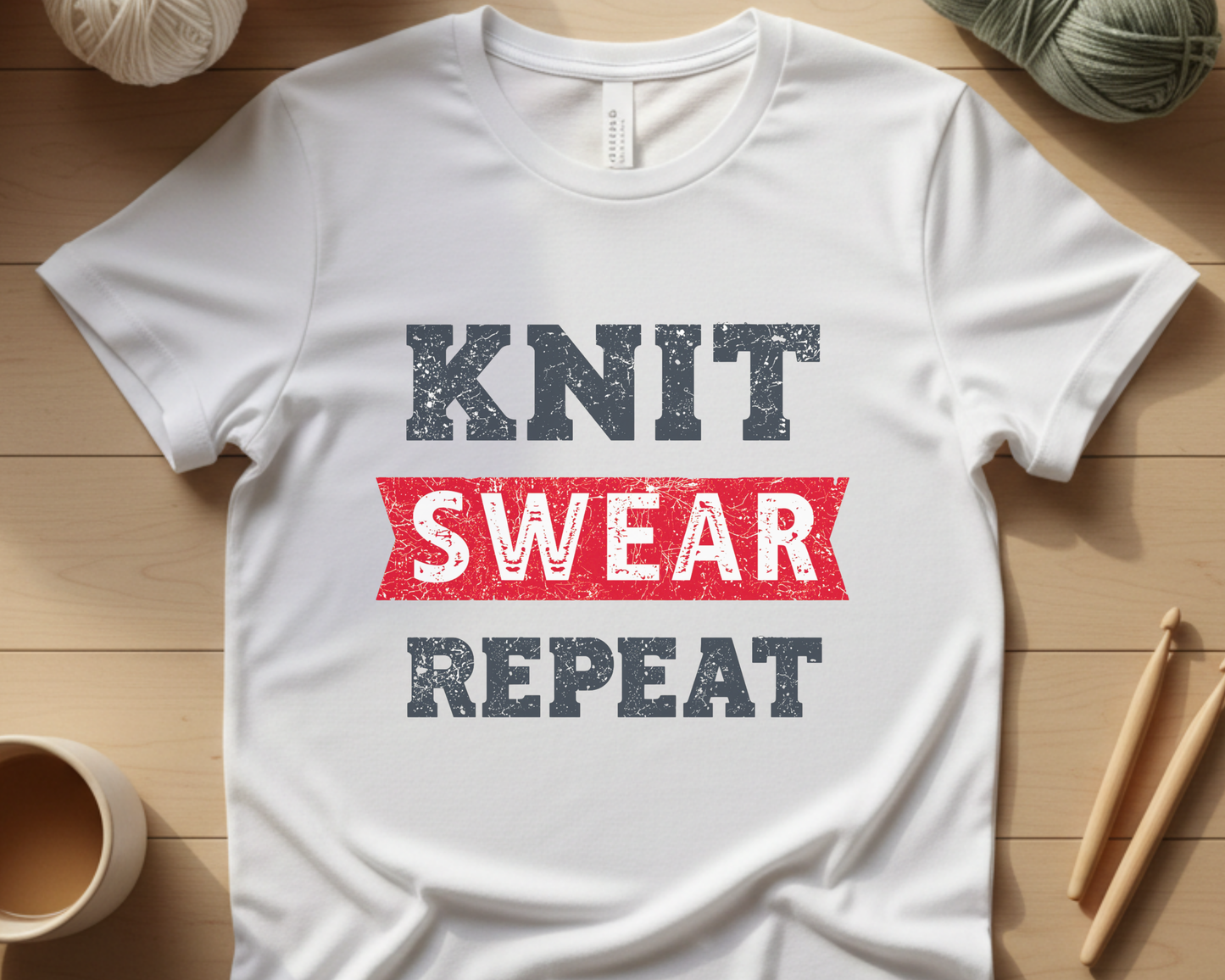 White t-shirt with 'KNIT SWEAR REPEAT' text on a wooden surface with yarn and knitting needles.