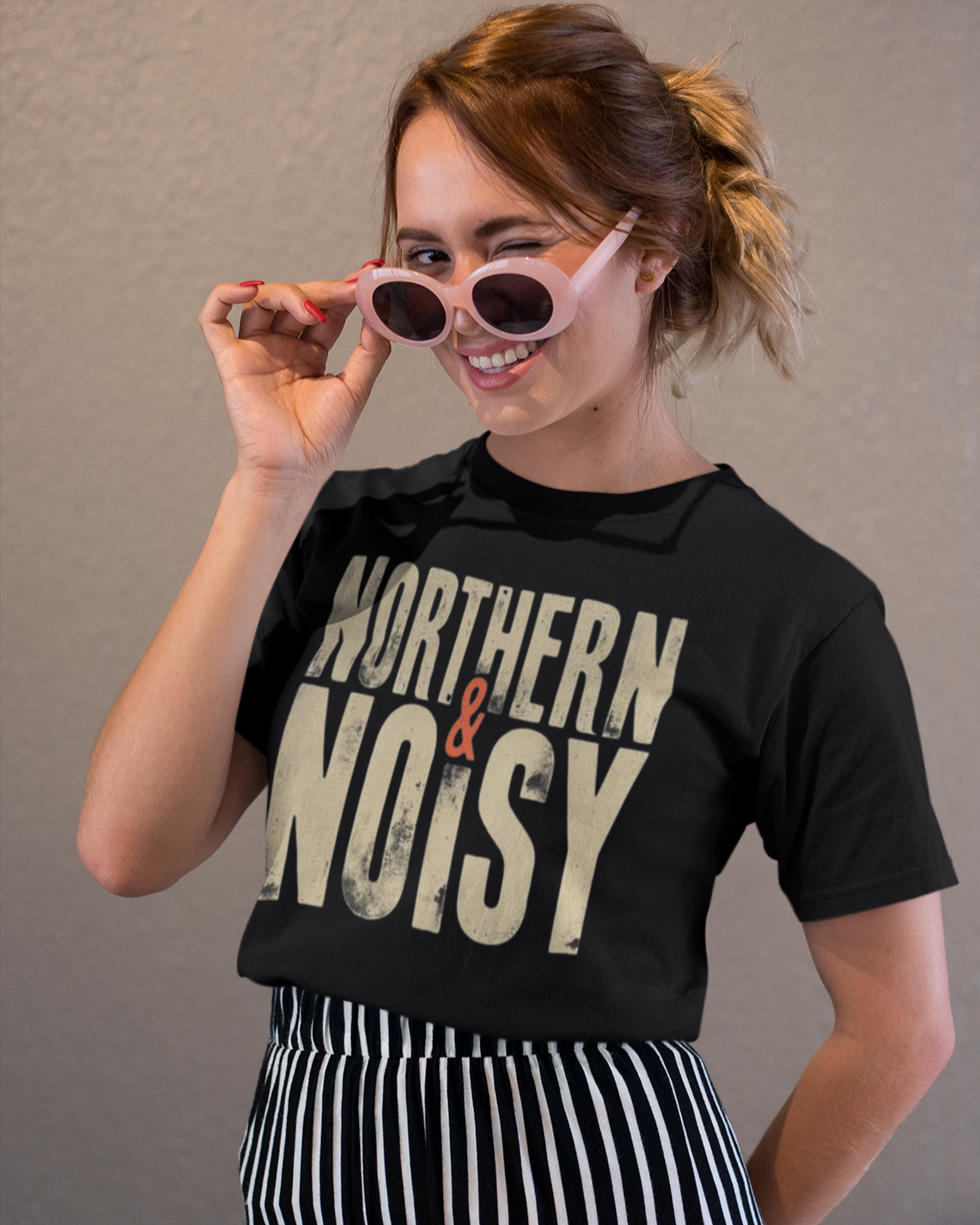 Woman wearing a black t-shirt with 'Northern & Noisy' text, holding sunglasses, against a plain background