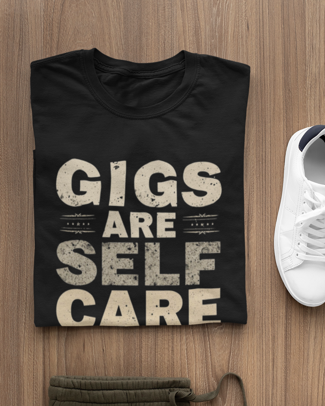 Black t-shirt with 'Gigs are Self Care' text on a wooden surface with a white sneaker.